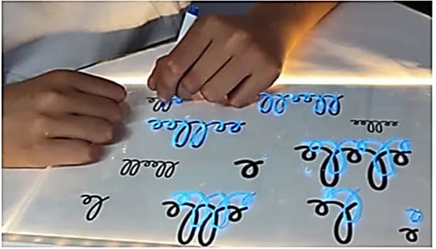 Deux mains d’enfant sur une table lumineuse, séries de boucles tracées au feutre fluorescent.