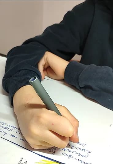 Main d’enfant écrivant au stylo, sur une feuille couverte de lignes manuscrites : la dictée de la lettre à l’ami.