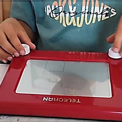 Mains d’enfant sur les deux molettes d’une ardoise magique Télécran rouge, un escalier tracé à l’écran.
