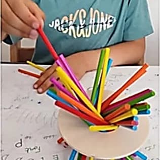 Main d’enfant posant un bâtonnet de couleur sur un fagot de bâtonnets plantés dans un socle rond en bois.