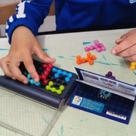 Mains d’enfant plaçant les pièces à billes colorées du casse-tête IQ Fit sur son plateau bleu, le boîtier ouvert à côté.