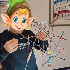 Enfant au visage remplacé par un lutin dessiné, posant une tige colorée sur le jeu d’équilibre Suspend, un enchevêtrement de fils métalliques.