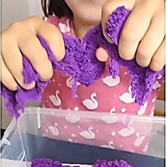 Mains d’enfant pétrissant du sable cinétique violet au-dessus d’un bac transparent.