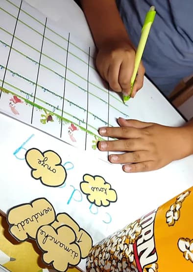 Main d’enfant au stylo vert sur une feuille de colonnes lignées, des mots découpés en forme de pop-corn et une boîte de pop-corn au premier plan.