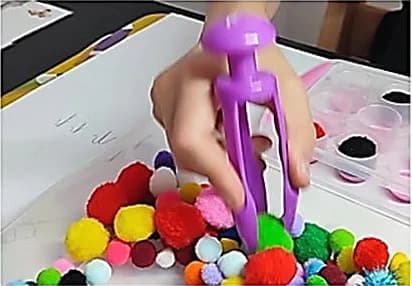 Main d’enfant tenant une grande pince violette au-dessus de pompons colorés et d’un bac à compartiments.