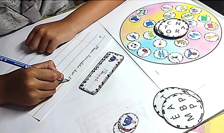 Main d’enfant écrivant au crayon sur une bande lignée, une roue de lettres et des jetons-lettres posés à côté.