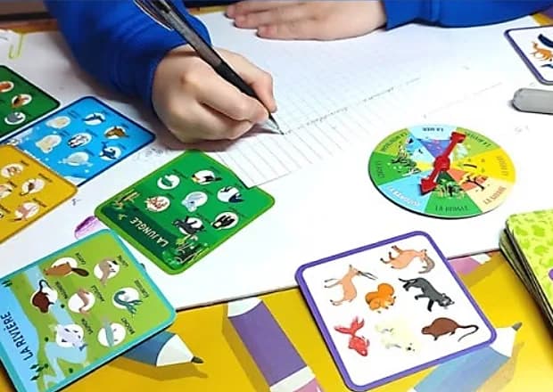 Main d’enfant écrivant au stylo sur un cahier quadrillé, entourée des cartes d’animaux et de la roue du jeu Speedo Memo.