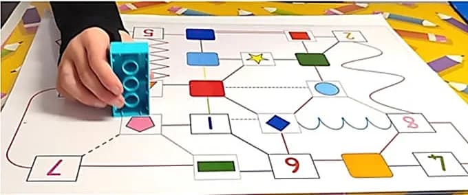 Une main d'enfant avance sa brique DUPLO sur un parcours codé de formes et de tracés