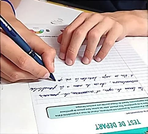 Main d’enfant écrivant au stylo bleu sur une feuille quadrillée intitulée « Test de départ », l’autre main posée à plat sur la feuille.